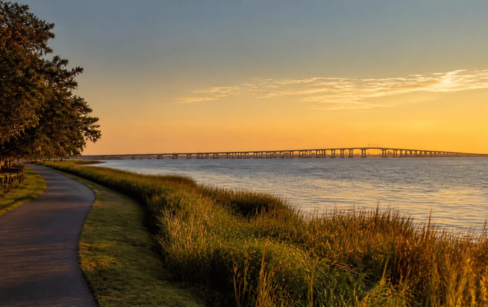 Hampton Roads waterfront walking path at sunset with marsh grass and a long bridge across the bay